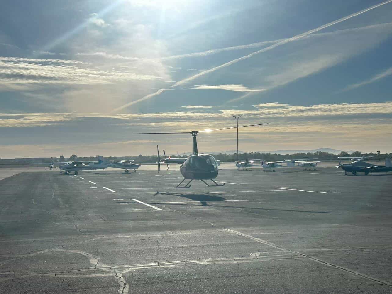 Robinson R44 on airport ramp at sunrise with fixed-wing aircraft in background